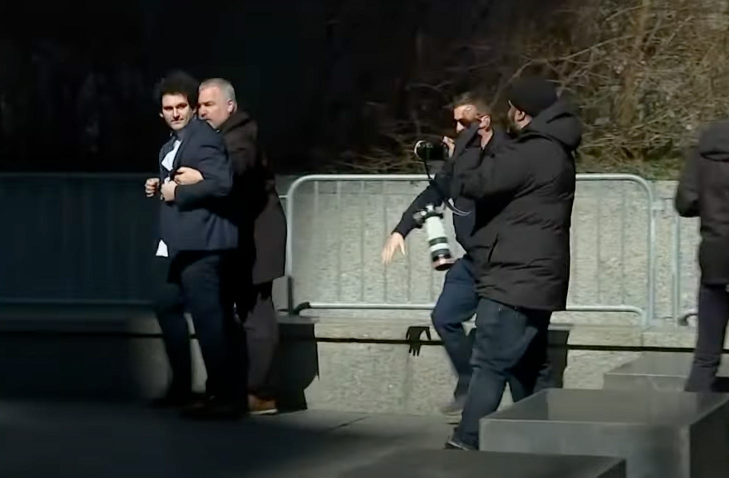 A bodyguard walks next to Sam Bankman-Fried with his arms underneath SBF's arms. They have just exited a crowd of photographers. SBF is wearing a navy suit and is looking back at the photographers while walking forwards. A bodyguard walks next to Sam Bankman-Fried with his arms underneath SBF's arms. They have just exited a crowd of photographers. SBF is wearing a navy suit and is looking back at the photographers while walking forwards.