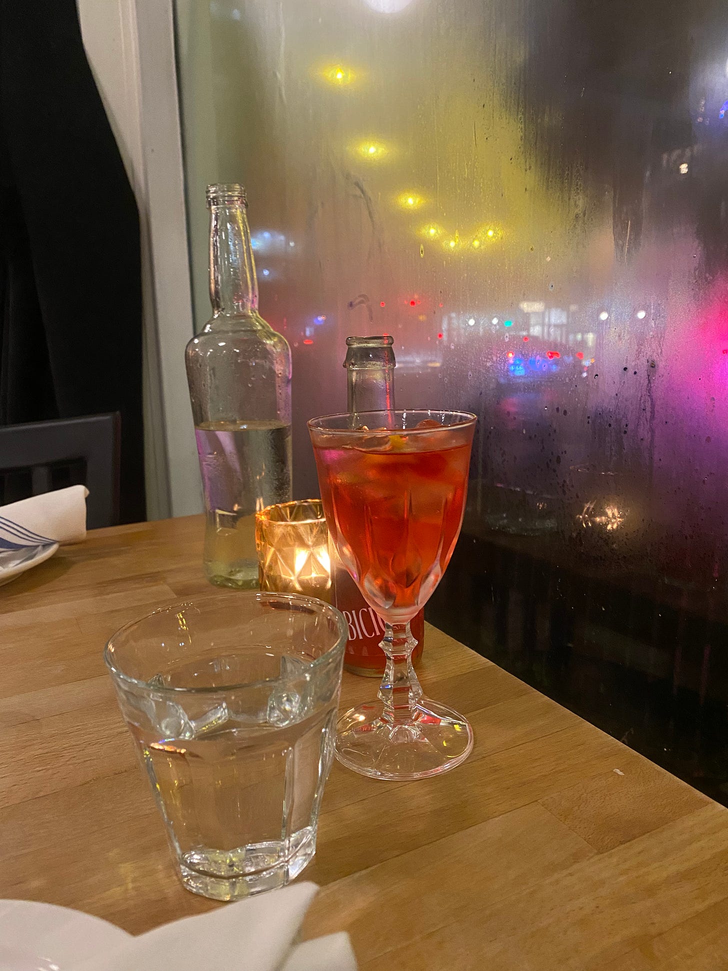 A table in a restaurant with glasses of water on it, and a crystal glass with a red drink and a slice of orange rind in it. There's a little bottle behind it that says 'bicicletta'. The window is foggy and it's dark out, and the lights from cars and the street outside are making the window light up in various colours.