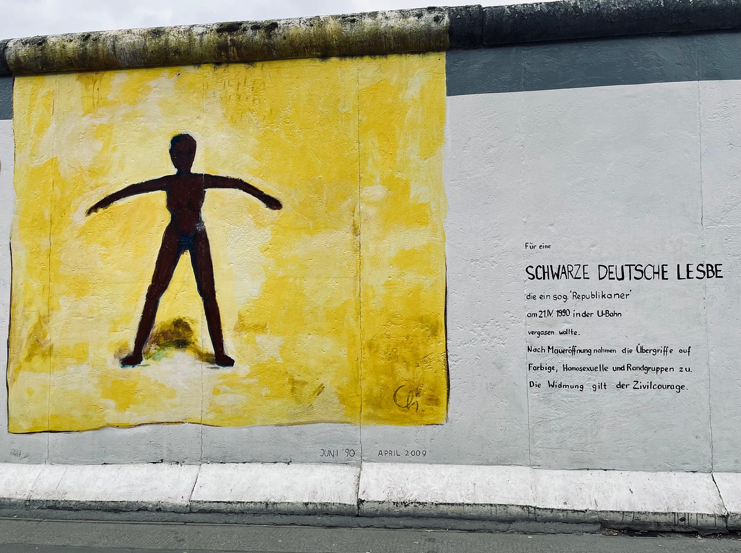 A mural on the Berlin Wall at East Side Gallery depicting a dark silhouette of a figure against a yellow background with arms outstretched. To the right, a text dedication reads: "For a Black German Lesbian who a so-called 'Republican' attempted to tear gas on 21.1.1990 in the subway. After the fall of the Wall, assaults on people of color, LGBTQ+, and marginalized groups increased. This dedication is for civil courage."