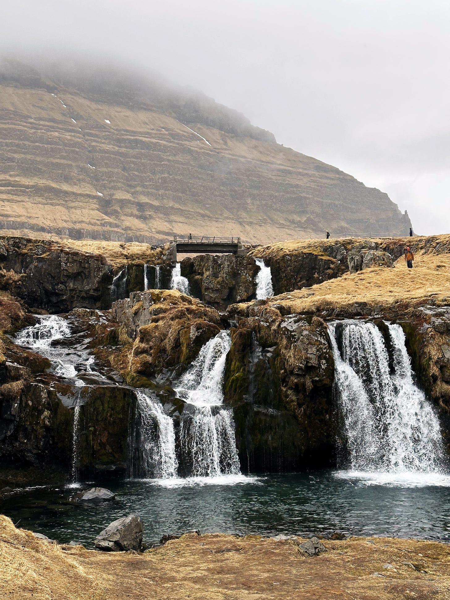 Kirkjufellsfossar, the cascading waterfalls in front of the mountain.  Shown against a large brown cliff with patches of snow people walk across a small bridge at the top of the falls.  The water flows into a pool below and eventually runs out to the sea on the right side of the frame.