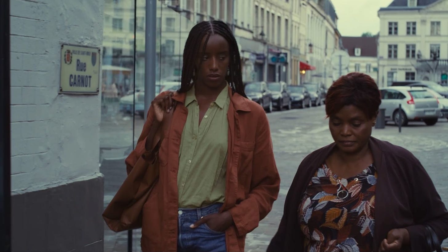 Two black women walk next to each other. One is younger and wearing braids down her back with a light green bkouse and camel colored jacket and bag. The elder Black woman is wearing a colorful shirt with blues and browns and a brown sweater. She is gazing down while the younger woman is gazing at her.