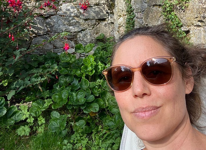 A selfie of me wearing sunglasses, with the background of my garden wall and some greenery.