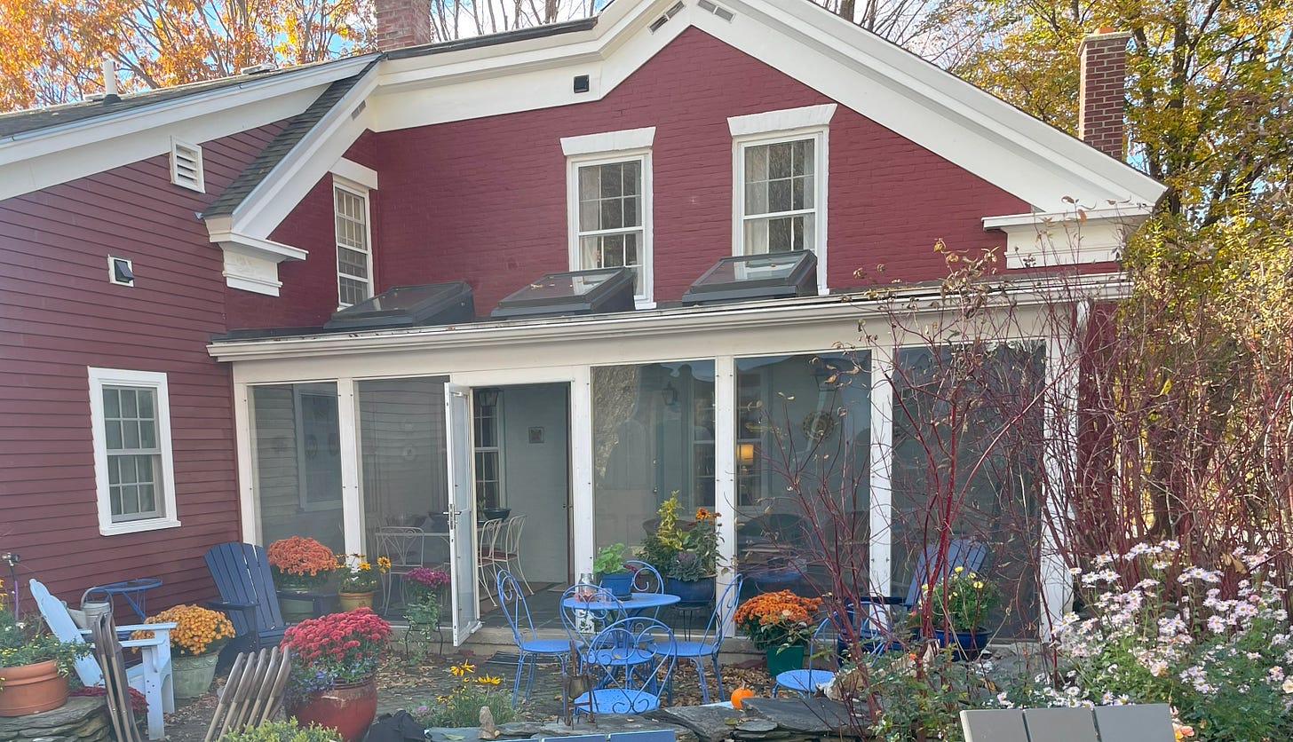 The backside of a red brick and clapboard house with a screened porch and then a patio in the foreground with potted mums and elegant garden furniture in a French blue. Fall leaves abound.