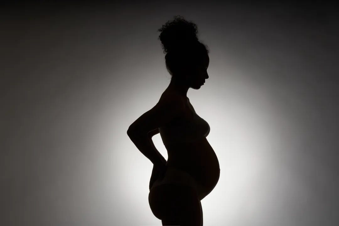 An image of the shadow of a pregnant person from the side, showing the thighs up. They have their arms behind them, hands braced on their lower back, and their hair is up.
