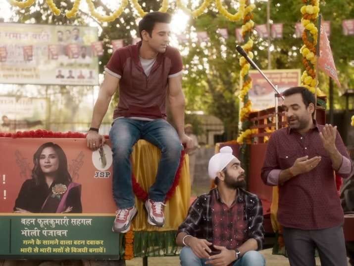 A still from Hindi film, ‘Fukr3y / Fukrey 3 ’. Hunny (Pulkit Samrat), Lali (Manjot Singh) and Panditji (Pankaj Tripathi) seem to be having a light conversation in an open environment with some flower decorations in the background. It seems like an election rally because of the poster in the background that has Bholi (Richa Chadha) in a black sari. The three men are all wearing a maroon, black white combination with jeans.