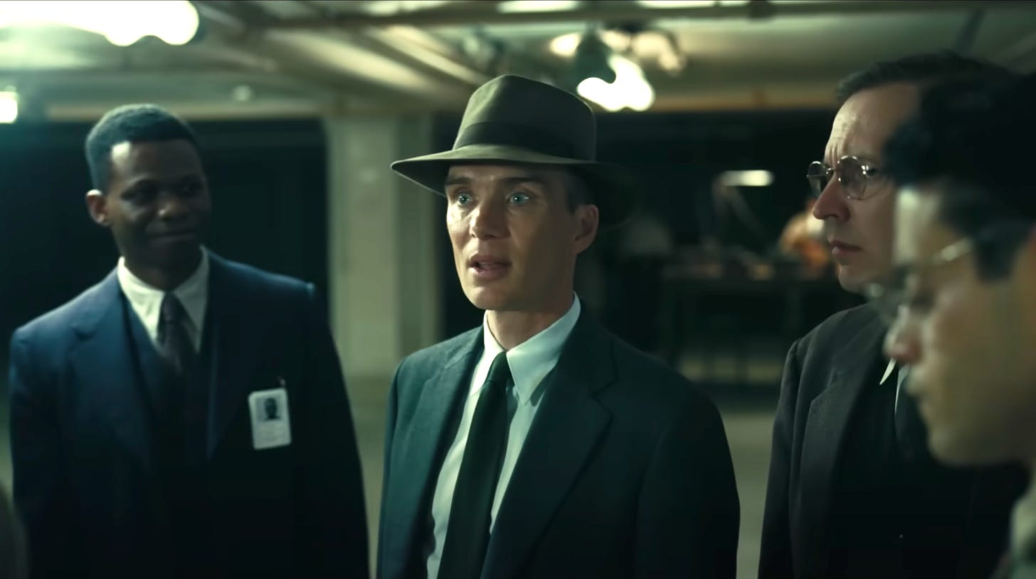 Murphy centered as Oppenheimer; Ronald Auguste as J Ernest Wilkins Jr slightly out of focus to picture left; Rami Malek out of focus in picture front right; I believe that is Ollii Haskivi as Edward Condon behind Malek