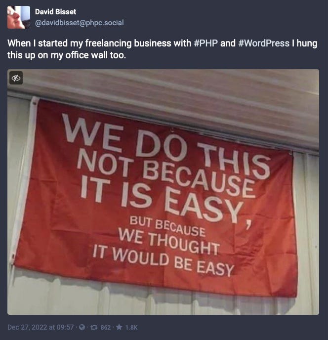 David Bisset Mastadon toot: When I started my freelancing business with #PHP and #WordPress I hung this up on my office wall too. Banner in picture reads: We do this not because it is easy, but because we thought it would be easy.