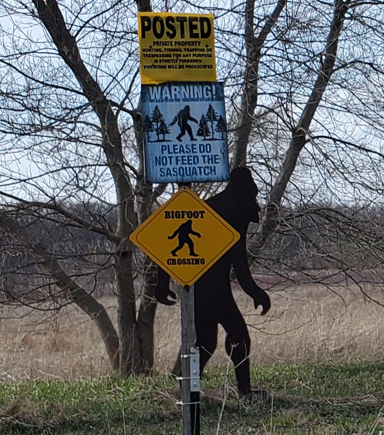 A private property marker, with two Bigfoot signs attached to it. One reads: “Warning! Please do not feed the Sasquatch.” The other says “Bigfoot Crossing.” Behind the sign is a silhouetted cut out of Bigfoot. 