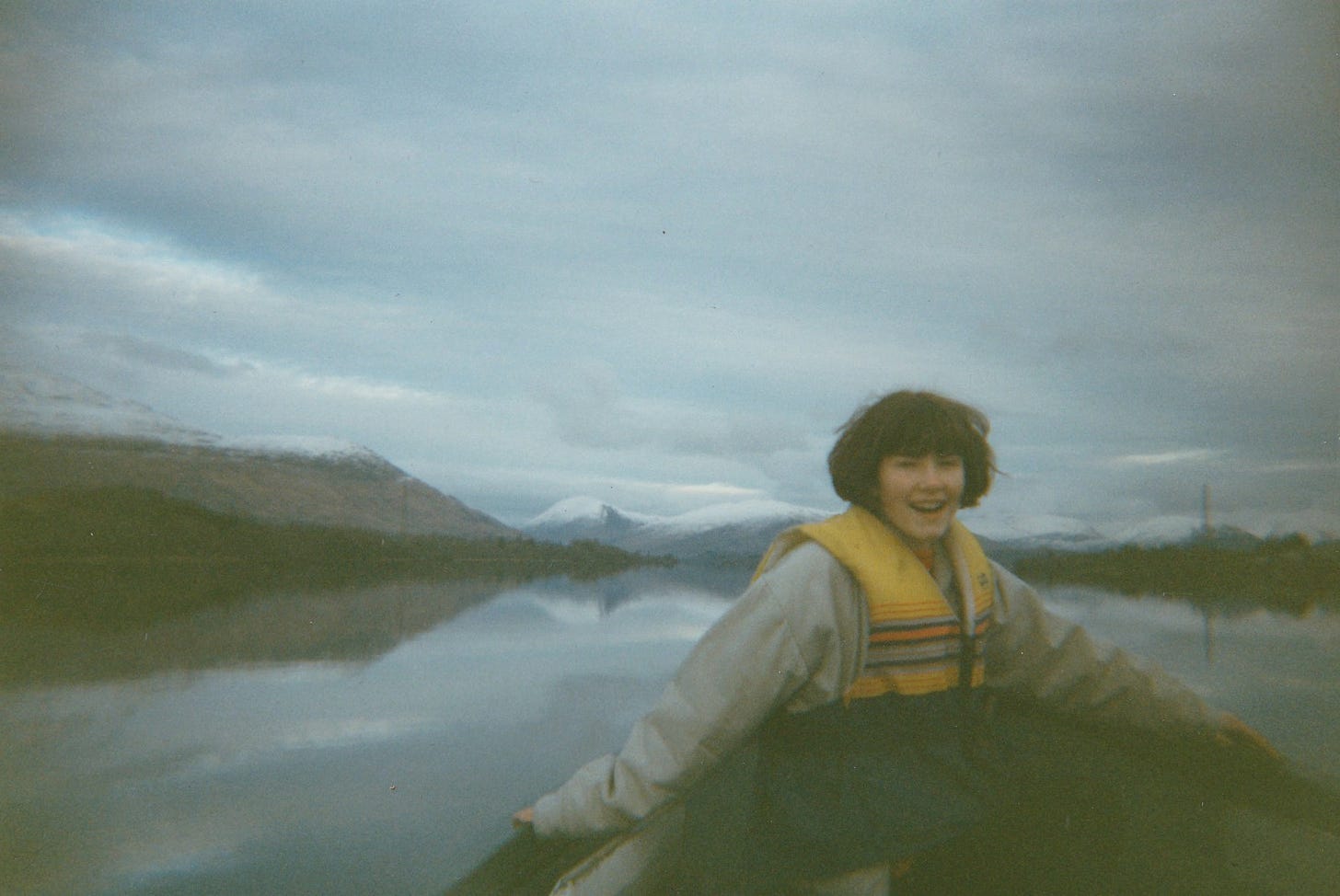 A girl in a yellow life jacket sitting in a small boat on a calm loch. Snowcapped hills in the distance.