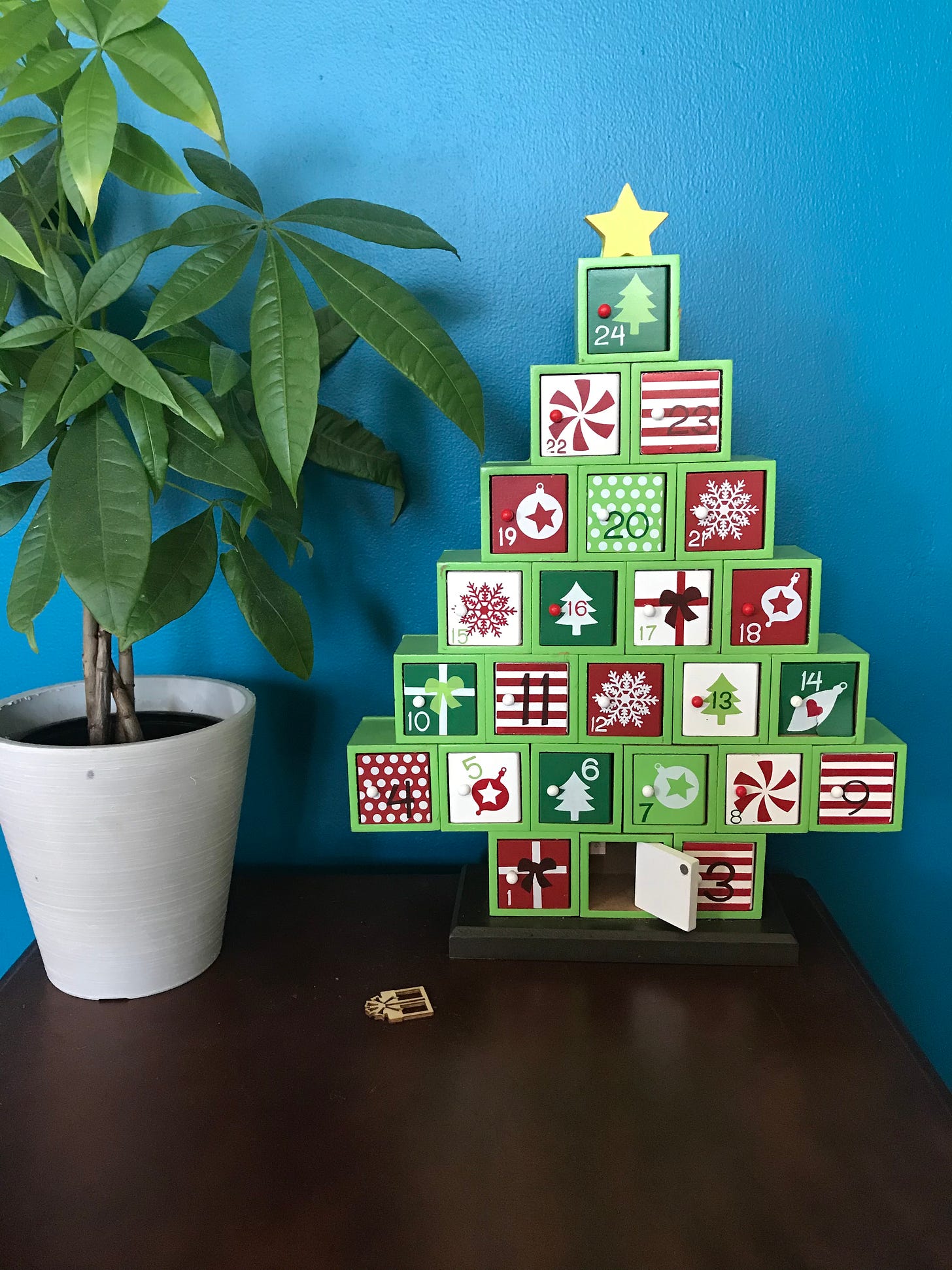 A wood table-top advent calendar in the shape of a Christmas tree. Small red and green doors open and climb up the tree to the top. Beside the advent calendar, a money tree. Behind both, a peacock blue wall.