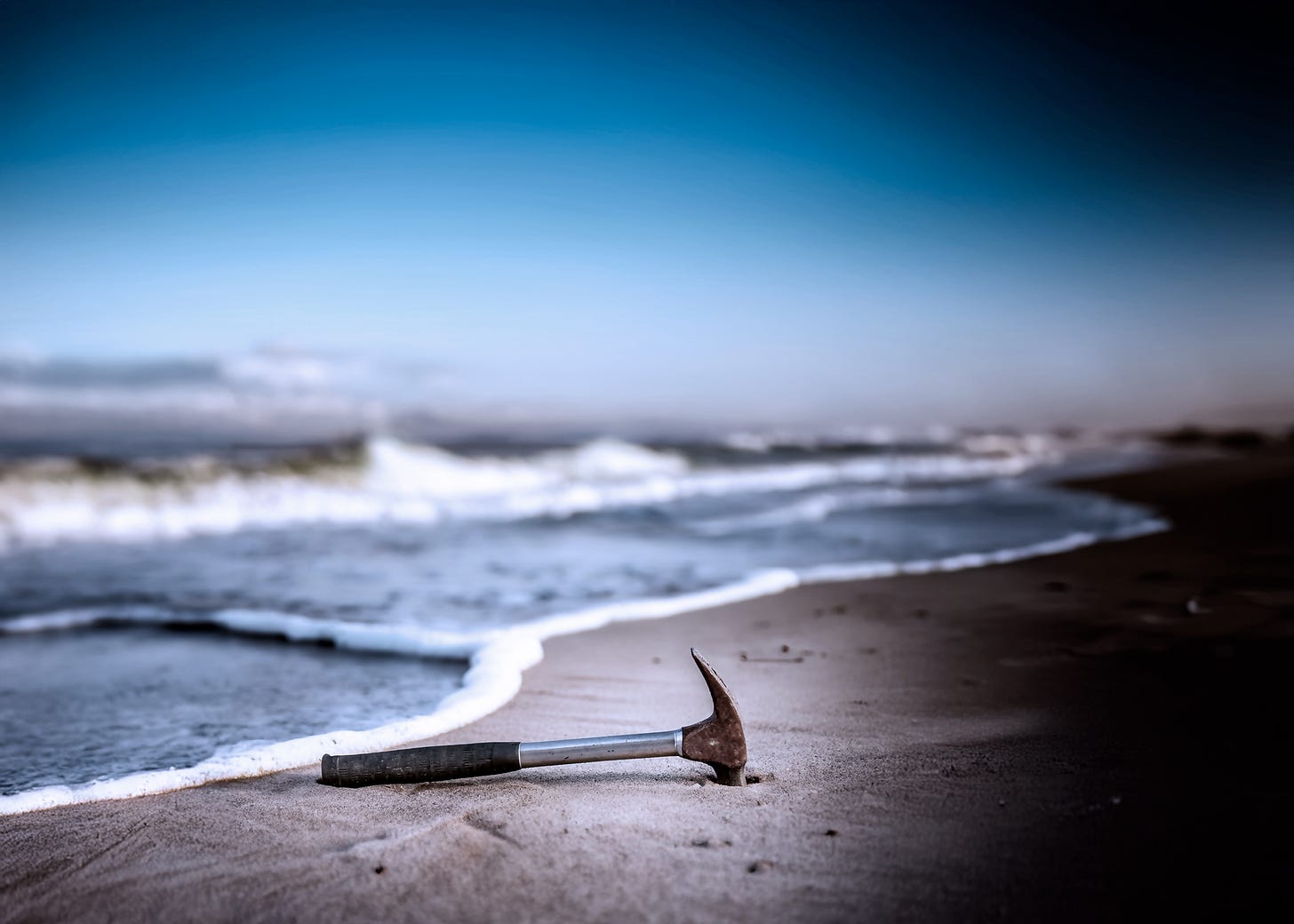 surreal image of a rusting hammer lying in the sand beside a stormy ocean with blackness creeping in from the side of frame