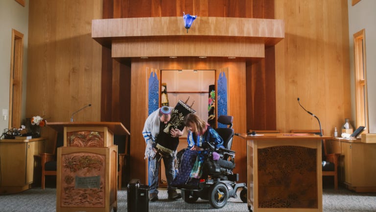 a synagogue sanctuary. On the bima are a man holding a Torah and a woman who uses a wheelchair