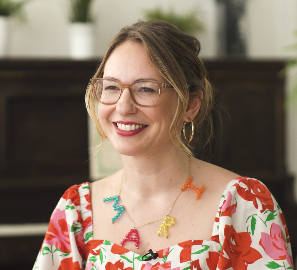 A women with light hair, brown glasses, and big hoop earrings smiles at something off camera. She is in a vibrant floral top and wears a colorful necklace that spells MARI.