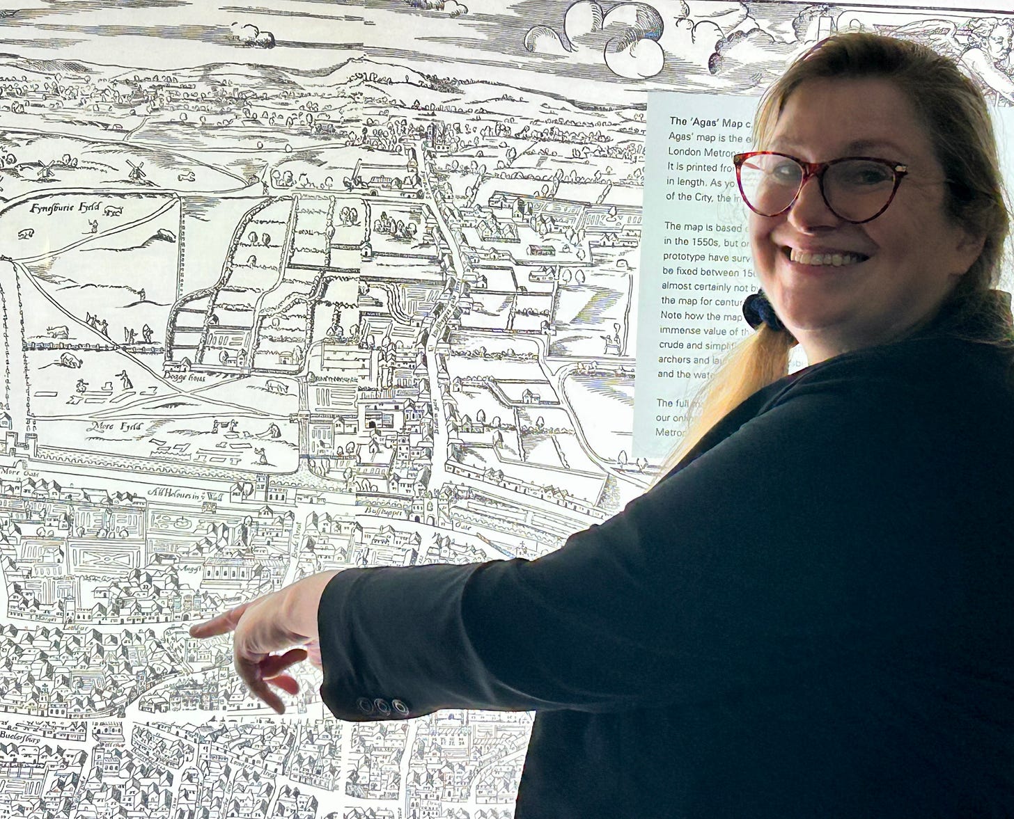 A woman wearing black is standing in front of an illuminated 16th century plan of London. She is pointing at a building with two towers at the front. 