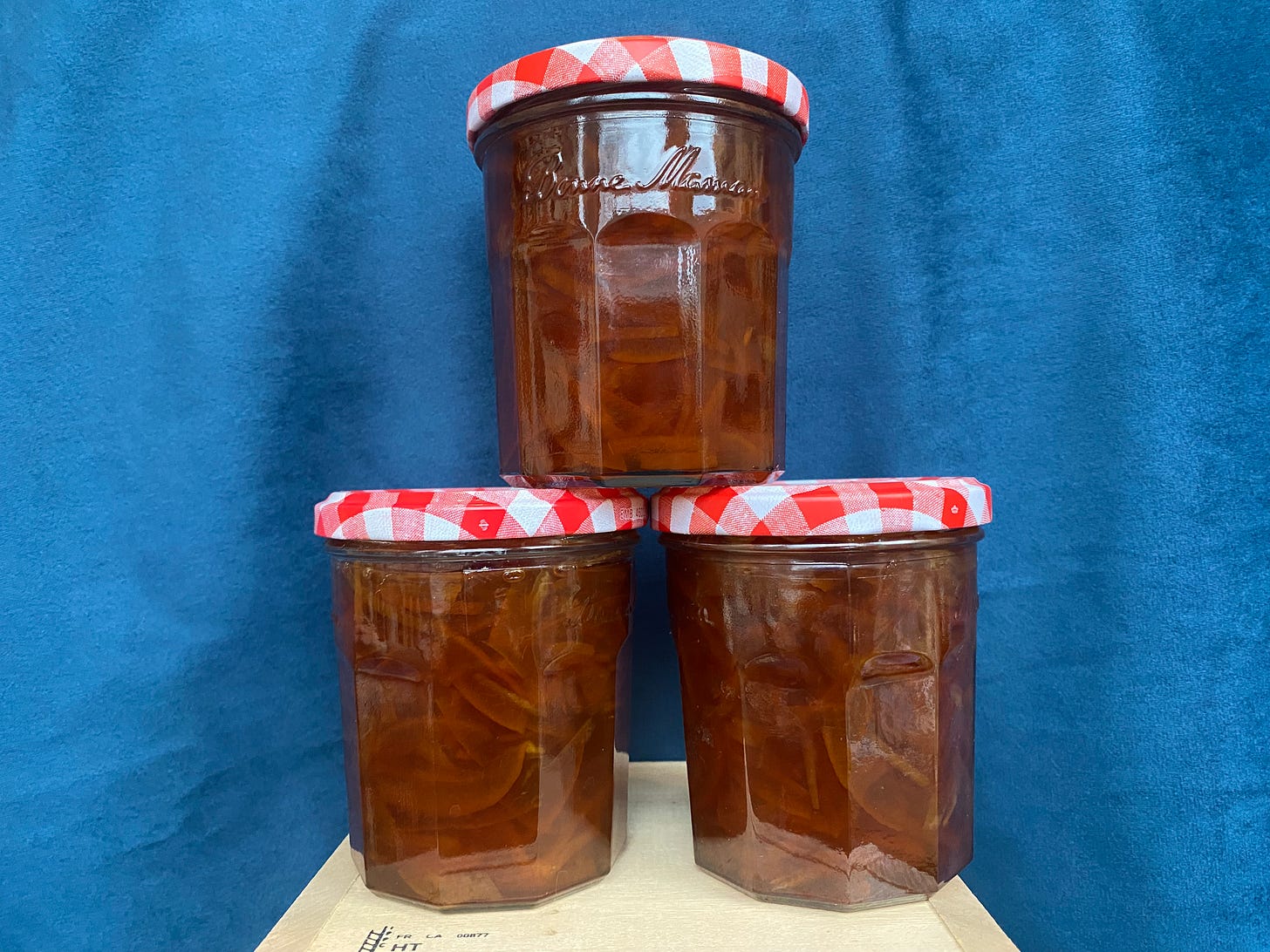 Three glass jars of deep orange marmalade, with the peel visible within, each with red and white jar tops. They are stacked on a wooden box in front of a green background.