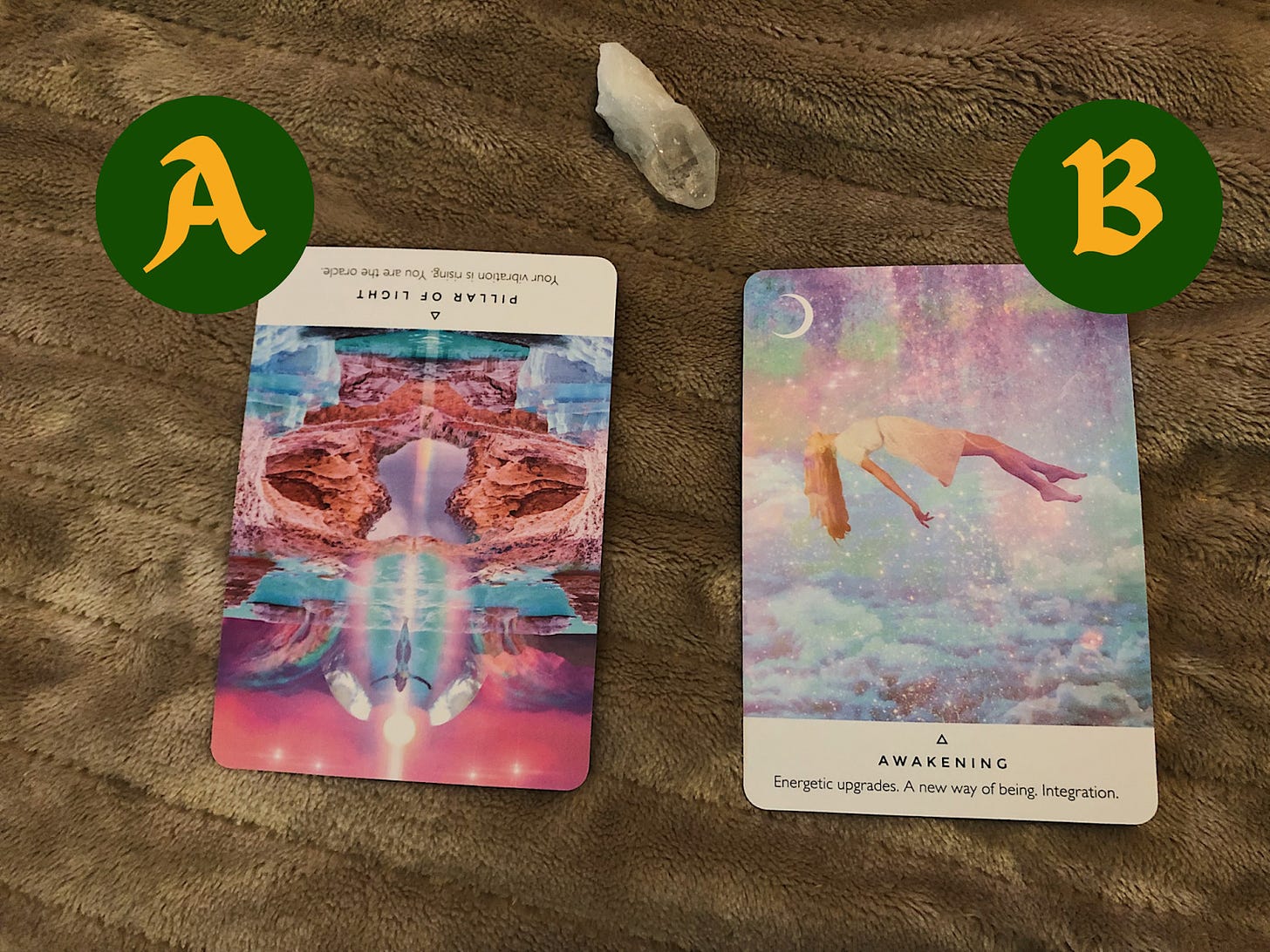 Two cards faced upright on a beige heated blanket with the left card being pink, purple, and blue with mountains and rivers and says "Pillar of light: Your vibration is rising. You are the oracle." The card on the right has a floating woman in a pastel purple and blue space and clouds that says "Awakening: Energetic upgrades. A new way of being. Integration."