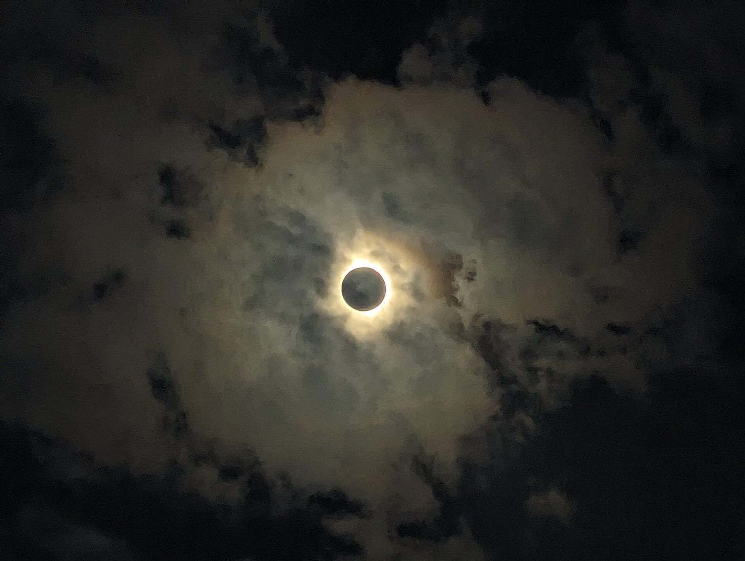 The eclipse photographed in a cloudy sky. In the center of the picture is a black circle ringed with yellow sunlight.
