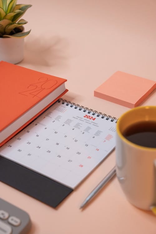 Free A peach-toned desk with 2024 planner, pen, coffee, and plant for organized productivity. Stock Photo