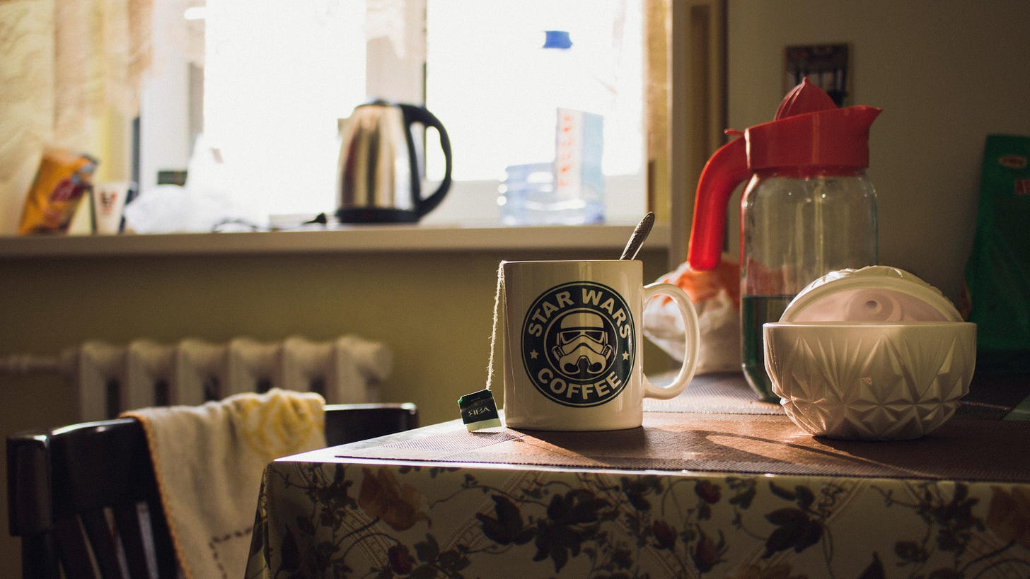 A photo of a domestic kitchen scene. There is a Star Wars mug on a table in the foreground. The mug has the string from a teabag hanging down its side. In the background, out of focus, a kettle is visible on a bright windowsill.