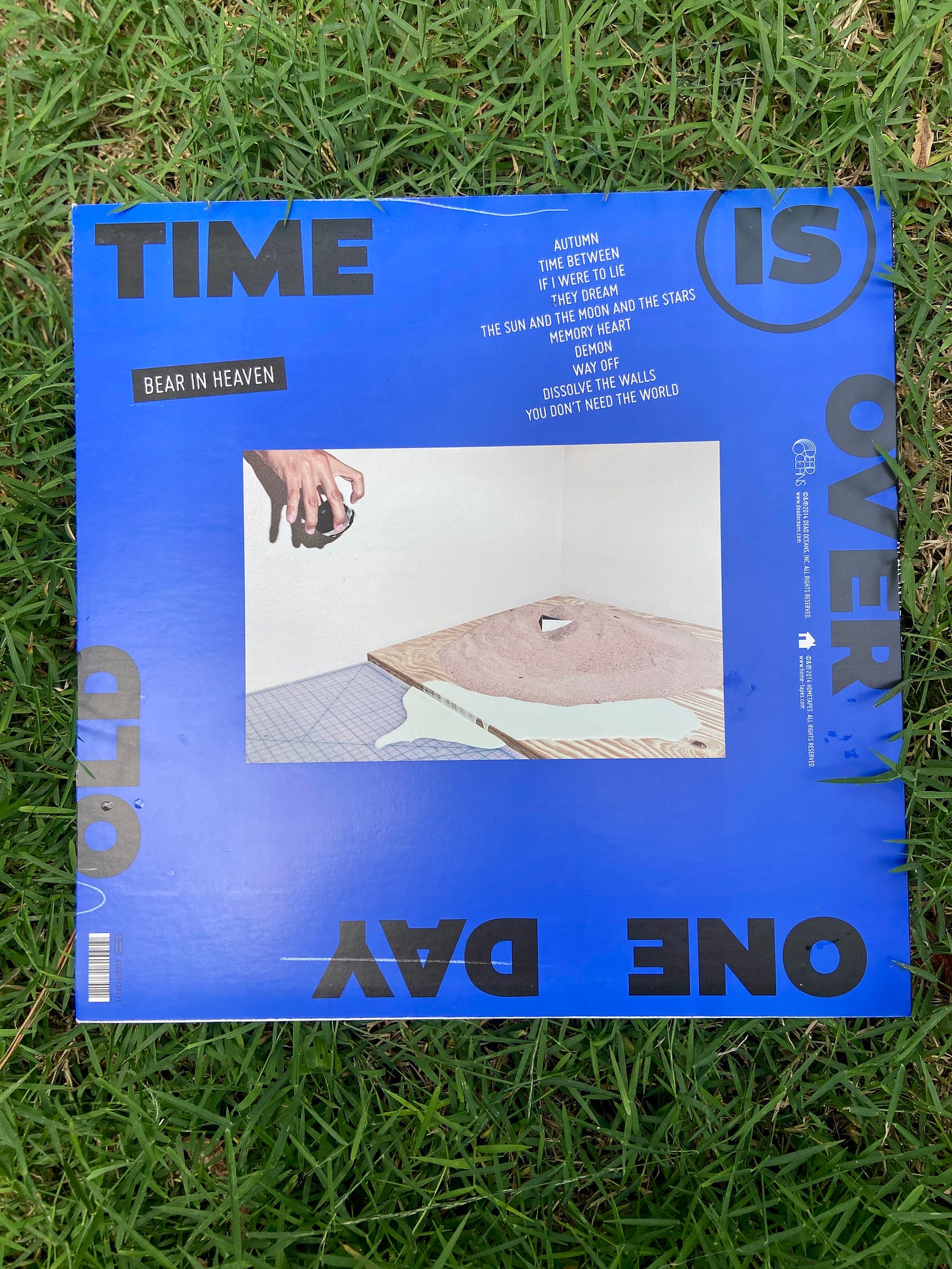 the back cover of Time is Over One Day Old by Bear in Heaven. it has a bright blue color, black and white text, and an image of a hand holding a canister over a wooden plank covered in a mound of sand and spilled paint.