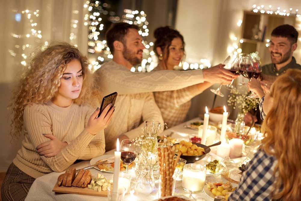 Young woman scrolling on her phone while the rest of the table celebrates a holiday cheers with win glasses.