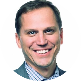 A professional headshot of a person wearing a gray suit jacket and blue checkered button-down shirt, smiling warmly at the camera against a white background.