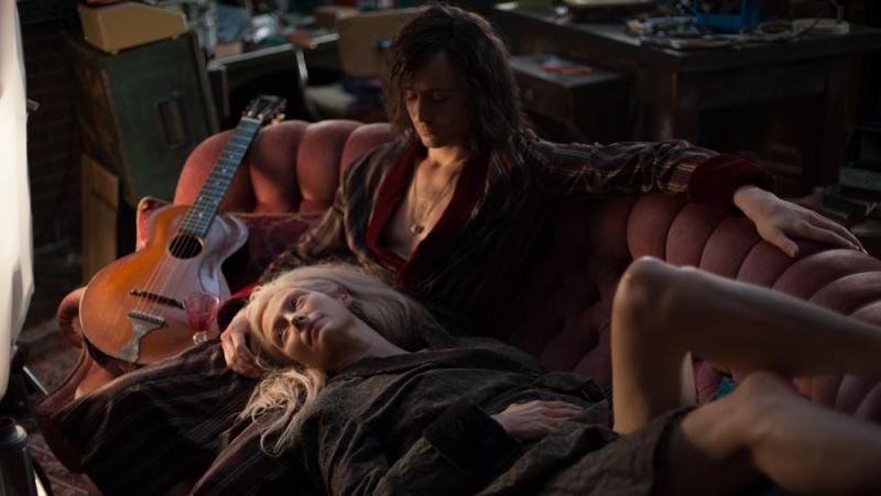 Eve deitada no colo de Adam, que está sentado em um sofá avermelhado com seu violão do lado.