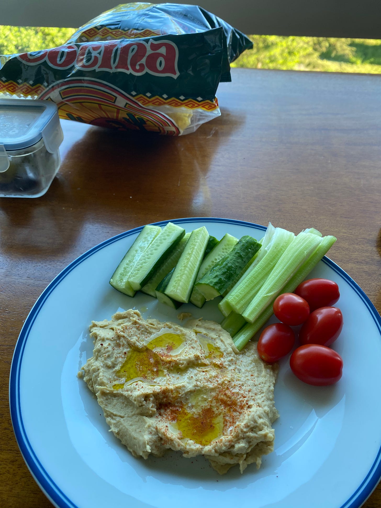 A white blue-rimmed plate with a smear of hummus drizzled with olive oil and paprika, surrounded on one side by grape tomatoes, celery, and cucumber sticks. In the background is an opened bag of La Cocina tortilla chips.