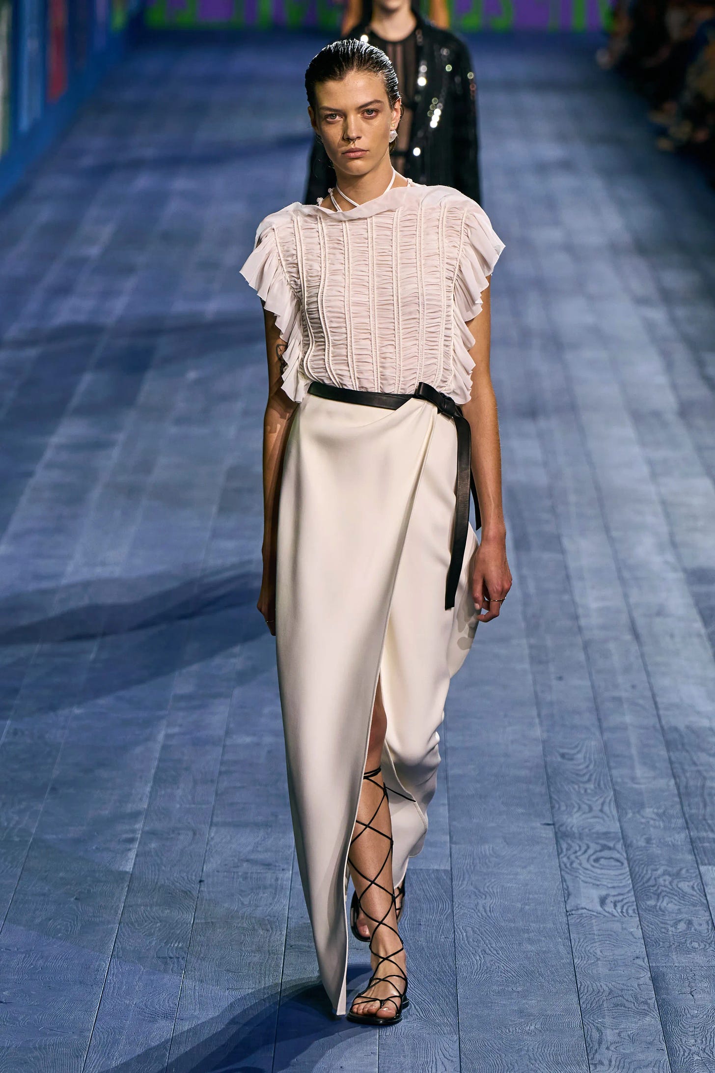 A female model walks a runway wearing a fluttering white top in horizontally pleated fabric paired with black-trimmed white wrap skirt and Roman-style lace-up sandals.