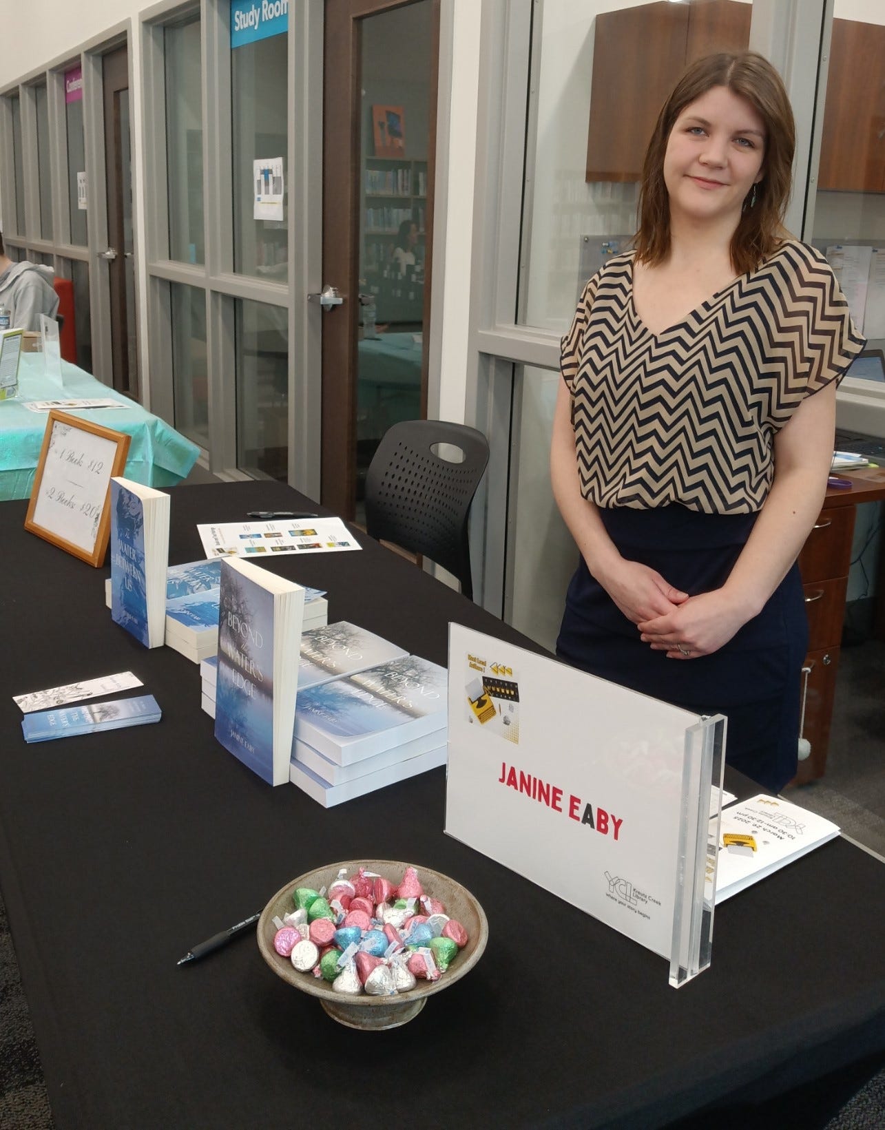 Janine Eaby at a local author event, standing at a table with her young adult portal fantasy books, Beyond the Water's Edge and The Water Between Us.