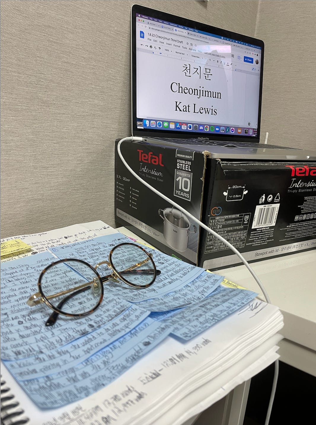 A photo of Kat’s atypically clean writing desk. Her computer is propped up on a box for a Tefal pot. The screen displays the title page for CheonJiMun. Next to her makeshift laptop stand is her printed manuscript. It’s opened to a page covered with lined sticky notes that are filled with her terrible handwriting. 
