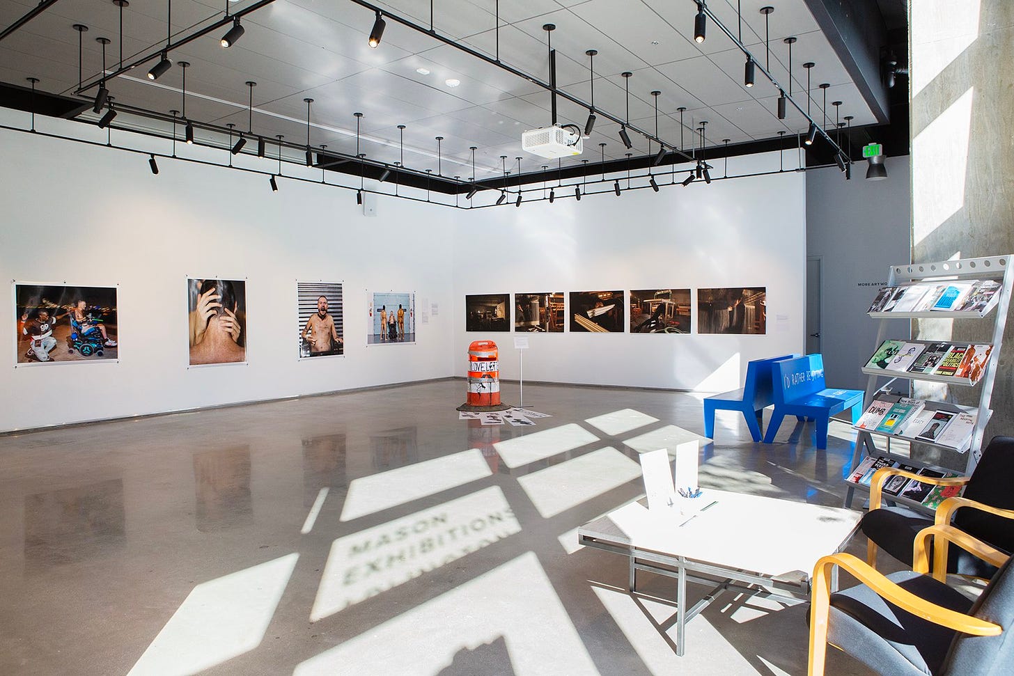 A gallery with photo works on a white wall, blue benches, and bright sunlight casting onto a cement floor.