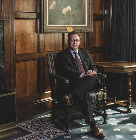 John Williams is seated in a wooded office wearing a suit with his hands clasped on his knee.