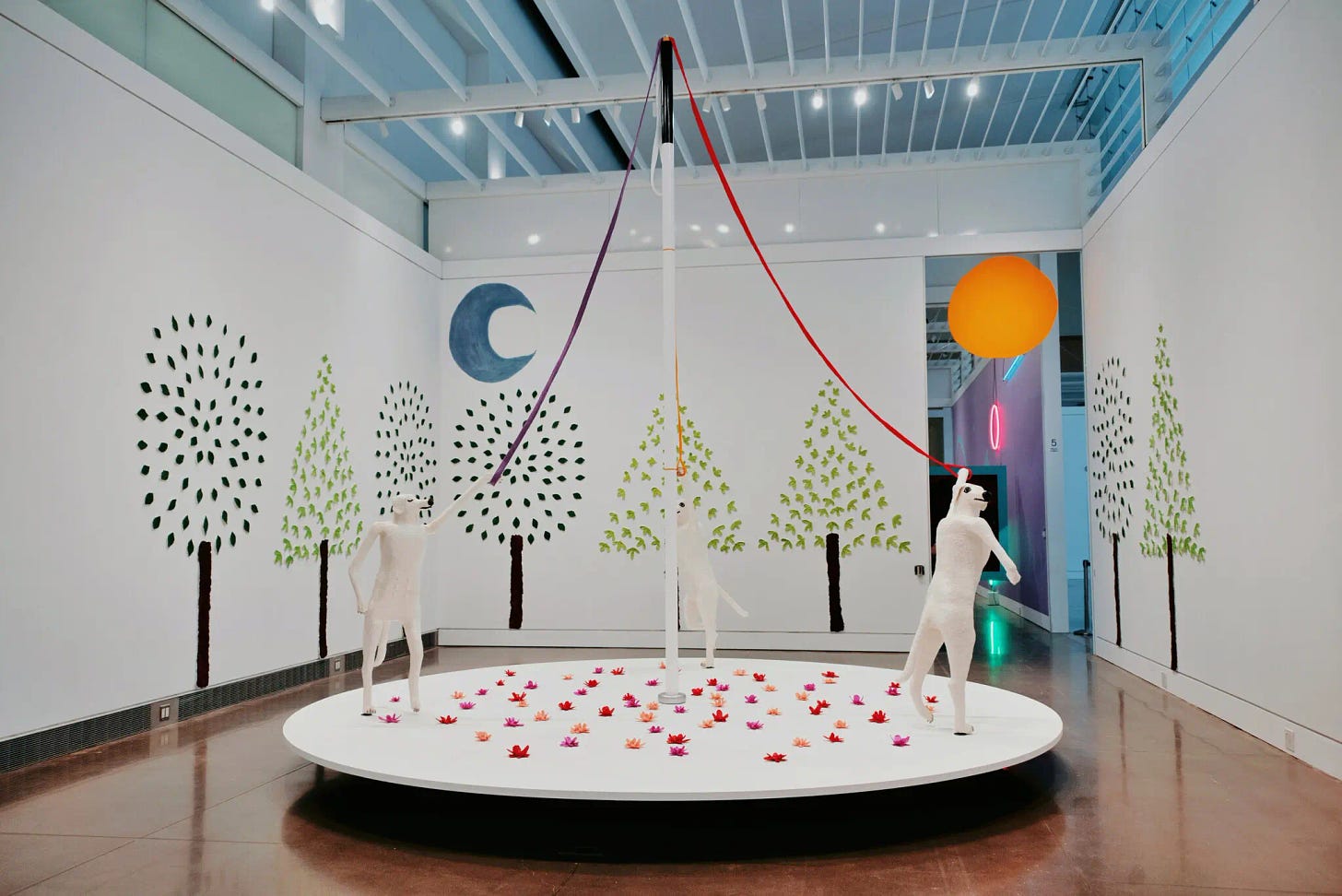 Three sculptures that are hybrids of the artist’s body and her dog’s body stand on hind legs, holding leashes, on a white platform in a gallery at the Queens Museum. There are art works on the walls resembling trees with round and triangular greenery.