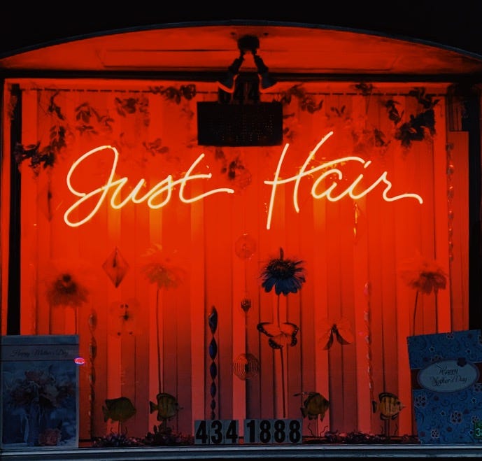 A photo of a store front with a neon sign in cursive reading “just hair”.