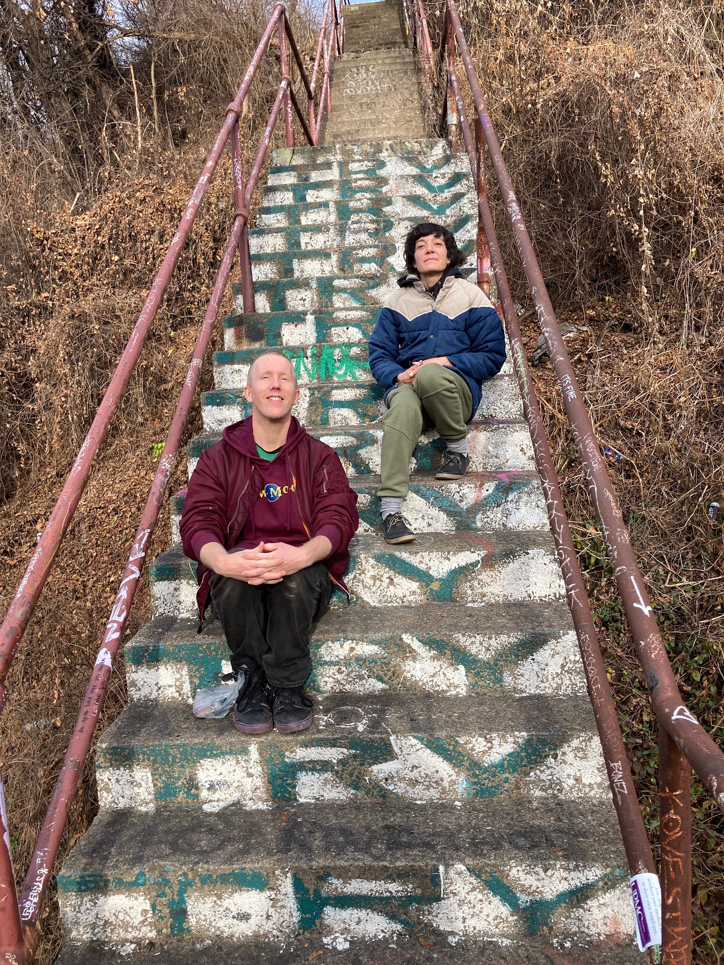 IMG DESCRIPTION: two white people sitting on a rusty run down set of concrete steps. The word "TRY" is painted on every step. They are surrounded by dead knotweed. They are smiling.