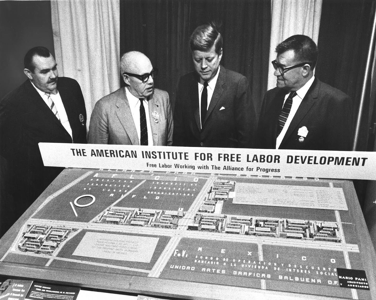 president JFK stands next to men in suits in front of a model of the american institute for free labor development