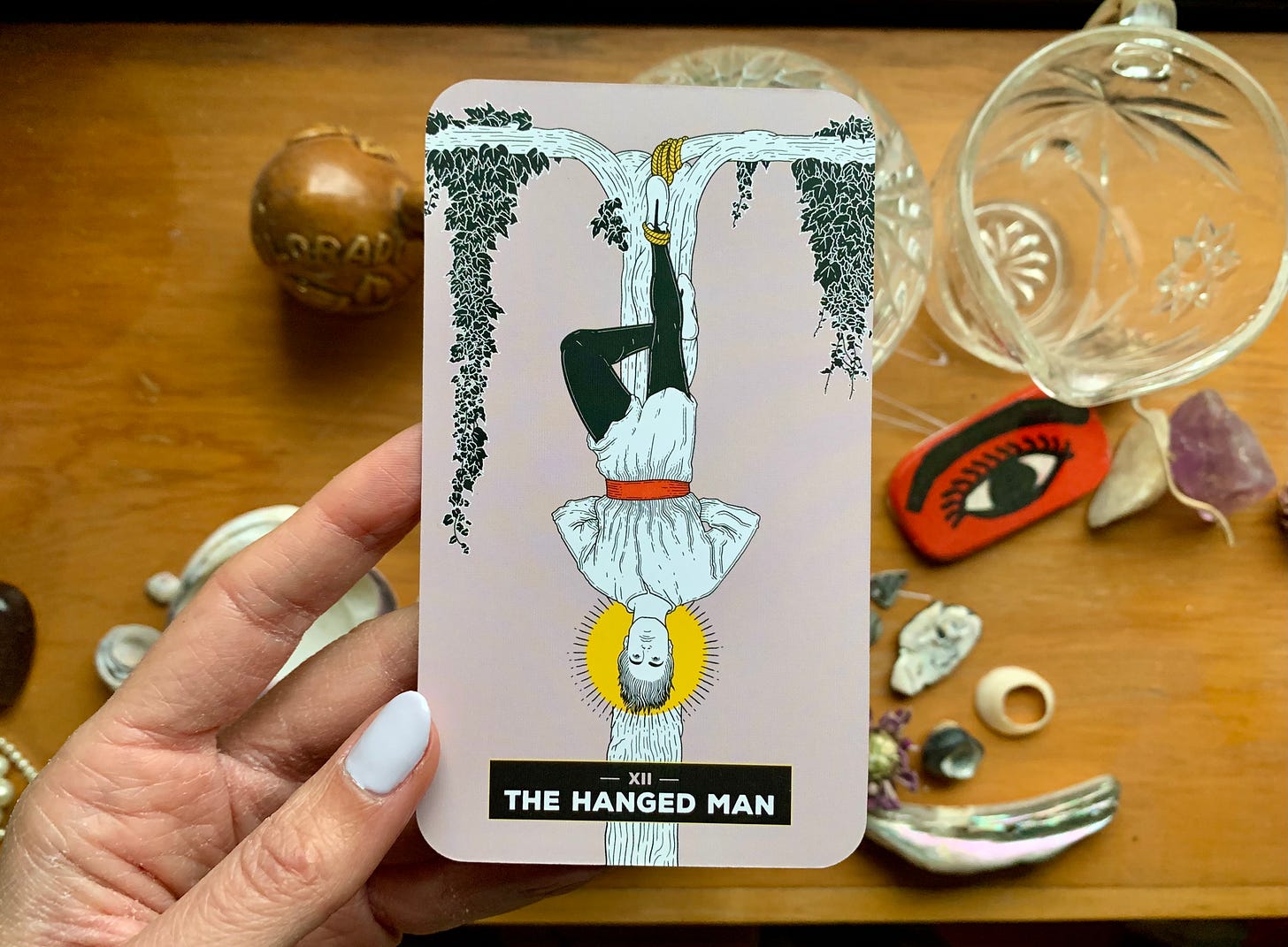 A hand is holding a tarot card, The Hanged Man by Xaviera Lopez from The Change Tarot. In the image a person is hanging from a tree by one foot with the other foot folded behind the straight leg. Their hands are folded behind their back and there is a halo around their head. Their facial expression appears to be restful.
