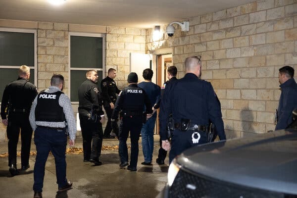 Luigi Mangione approaches a lighted door, wearing handcuffs and surrounded by police officers.