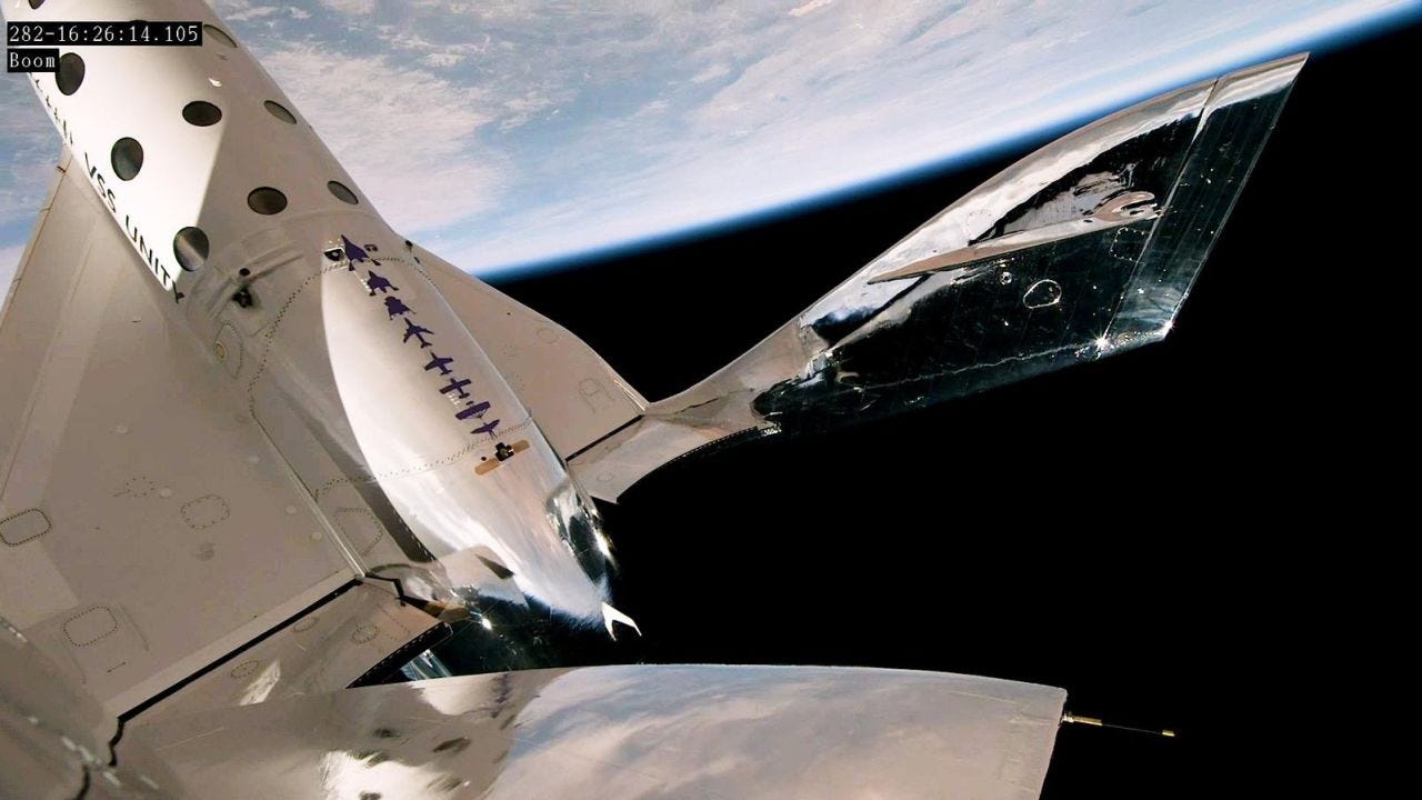 The image shows a white spaceplane named VSS Unity against a small portion of the Earth. The spaceplane has a sleek and elongated shape with a rounded nose and two wings extending outward. It is attached to a larger carrier aircraft called VMS Eve. Unity 25 and VMS Eve are flying in close formation, with Unity 25 positioned beneath the wings of VMS Eve. The sunlight illuminates the spacecraft, casting a shadow on the carrier aircraft. The scene conveys a sense of motion and excitement, capturing Virgin Galactic's test flight to suborbital space.
