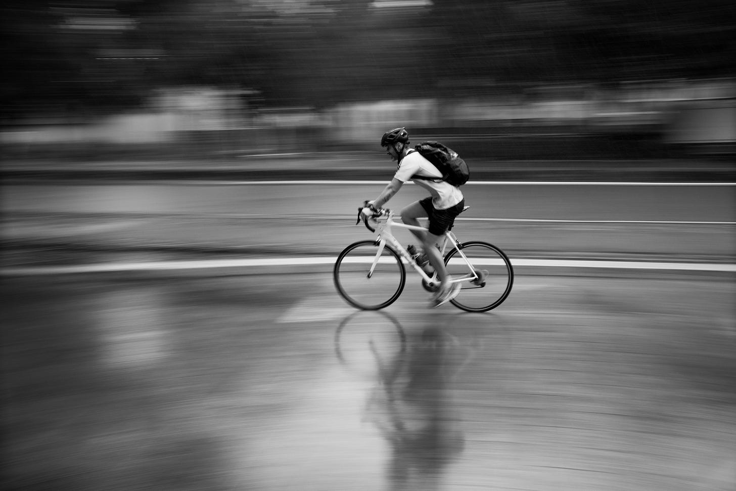 A black-and-white image of a commuter on a white road bike peddling furiously through the rain.
