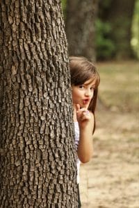 A cute little girl is hiding behind a tree and peeks around holding her finger to her lips saying, shhh