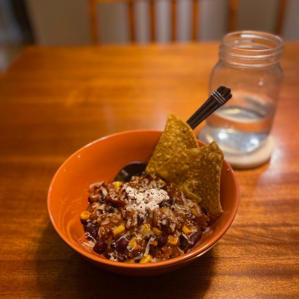An orange bowl of chili with corn, topped with grated cheese, sour cream, and a few tortilla chips. A glass of water sits on a coaster behind it.