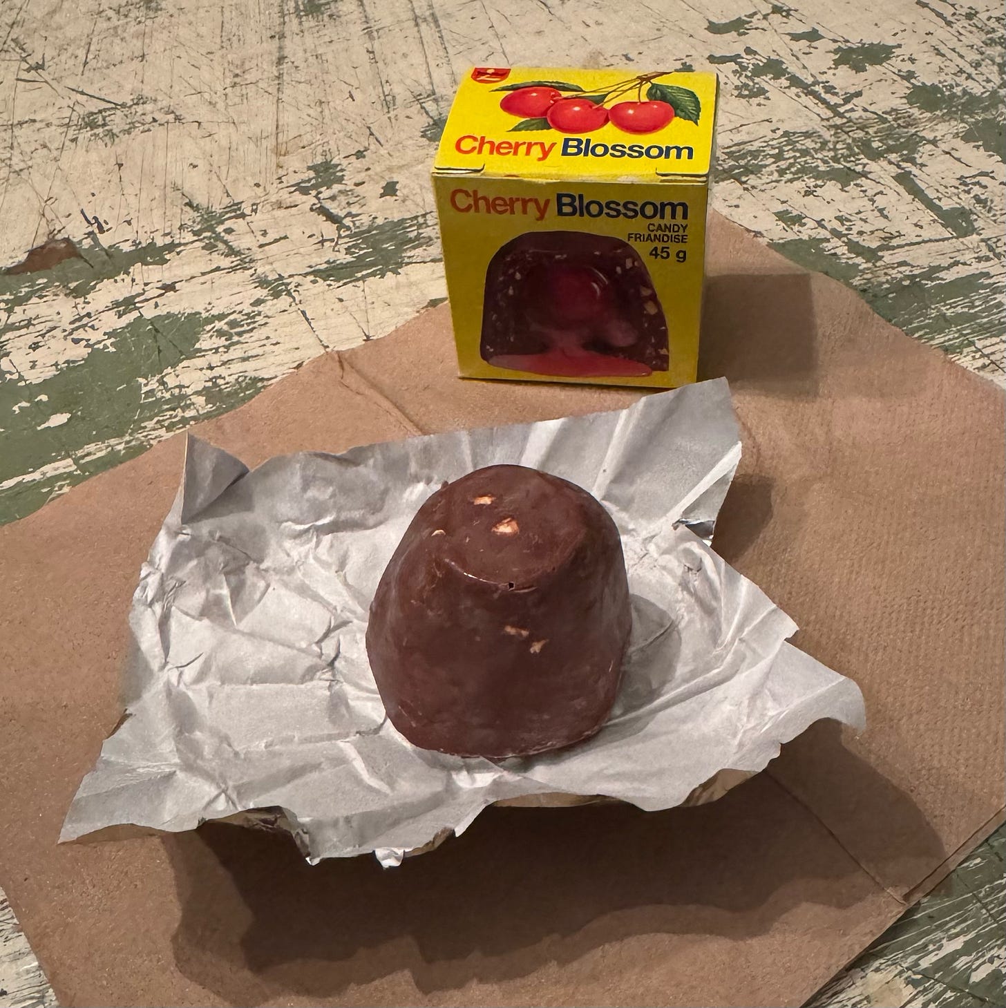 An unwrapped Cherry Blossom candy sitting on its opened foil wrapper, which is on top of a napkin. The Cherry Blossom box is behind it. The front of the box shows a cross section of the candy, with a cherry in the middle and poink goo pouring out around it. On the top of the box there are three cherries attached by stems and a few leaves.