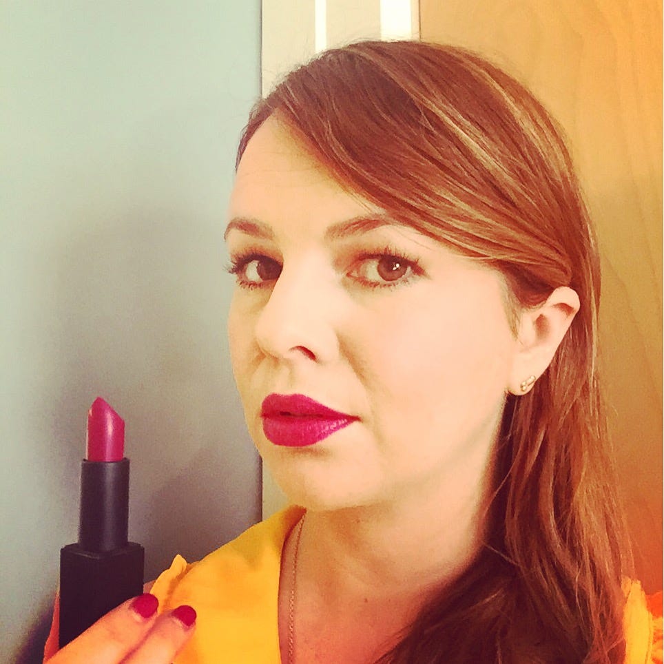 Amber holds up an open lipstick that matches the shade on her lips. She looks into camera.