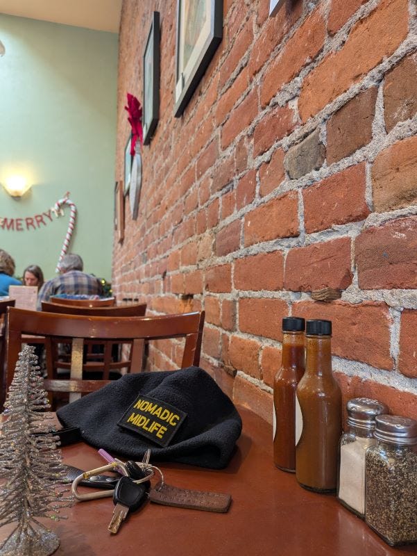 a hat and keys, plus bottles of hot sauce, salt and pepper shakers, and a little silver Christmas tree decoration on a wooden cafe table against a red brick wall covered with framed art. Another table with restaurant patrons is in the background.