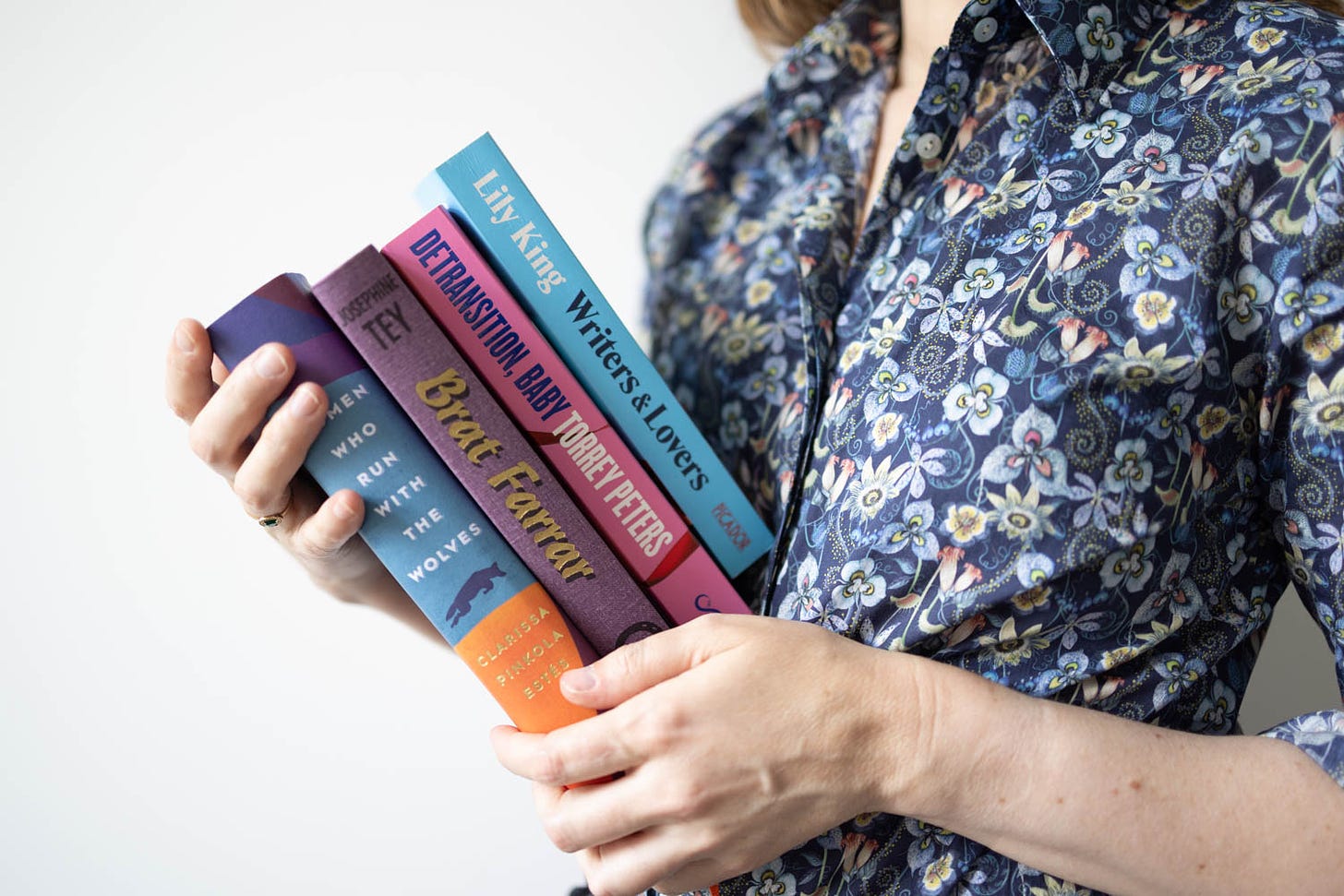 White woman in blue floral blouse holds a stack of books: Women who run with the wolves, Brat Farrar, Detransition Baby and Writers & Lovers