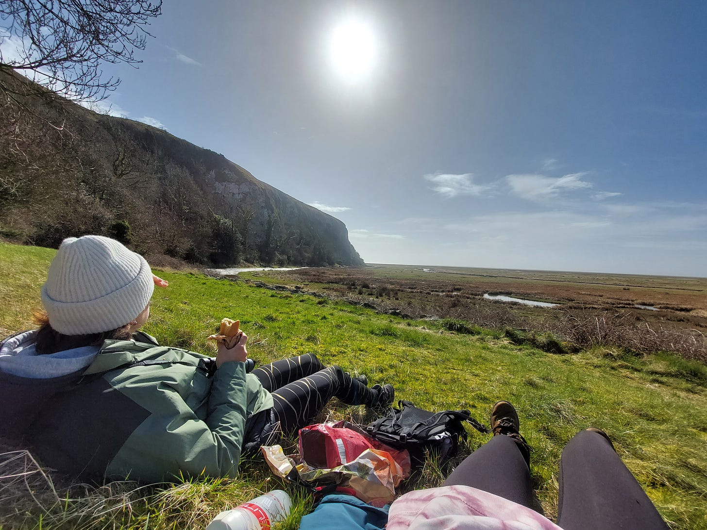 One person reclining eating a sandwich whilst looking out at a spectacular view. A cliff to the left, blue sky and marsh land. The legs of the photographer are in shot too. The best picnic spot!