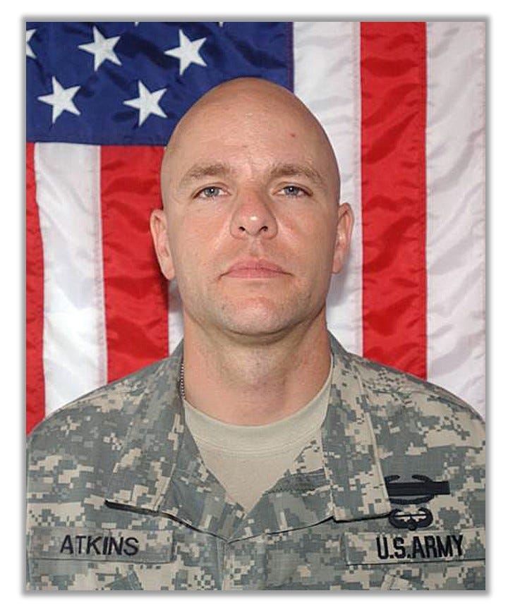 Headshot of Travis Atkins in his fatigues. A flag is in the background. 