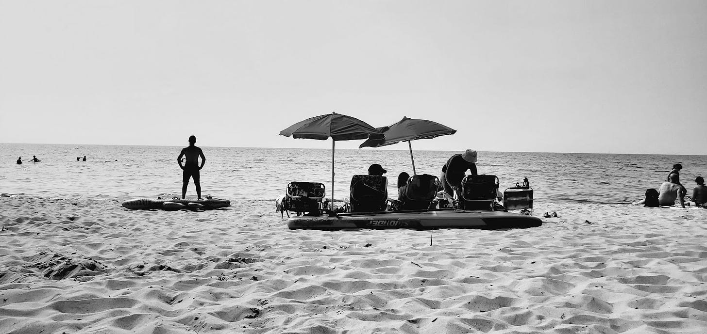 A black-and-white photo of people with chairs and umbrellas at the beach.